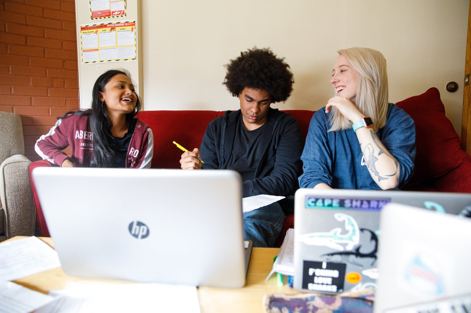 three students sitting at a couch laughing and studying, with laptops resting on a table in front of them