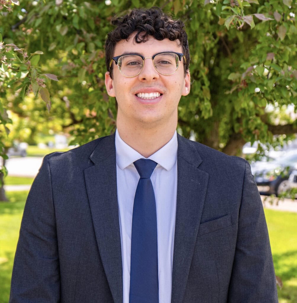 A man with short curly brown hair wearing a blue suit and tie smiling at the camera. 