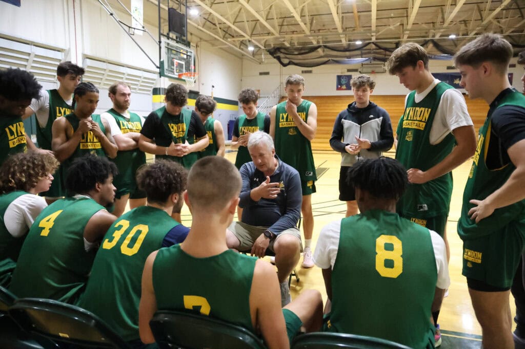 A group of VTSU Lyndon men's basketball players standing in a circle around their coach.