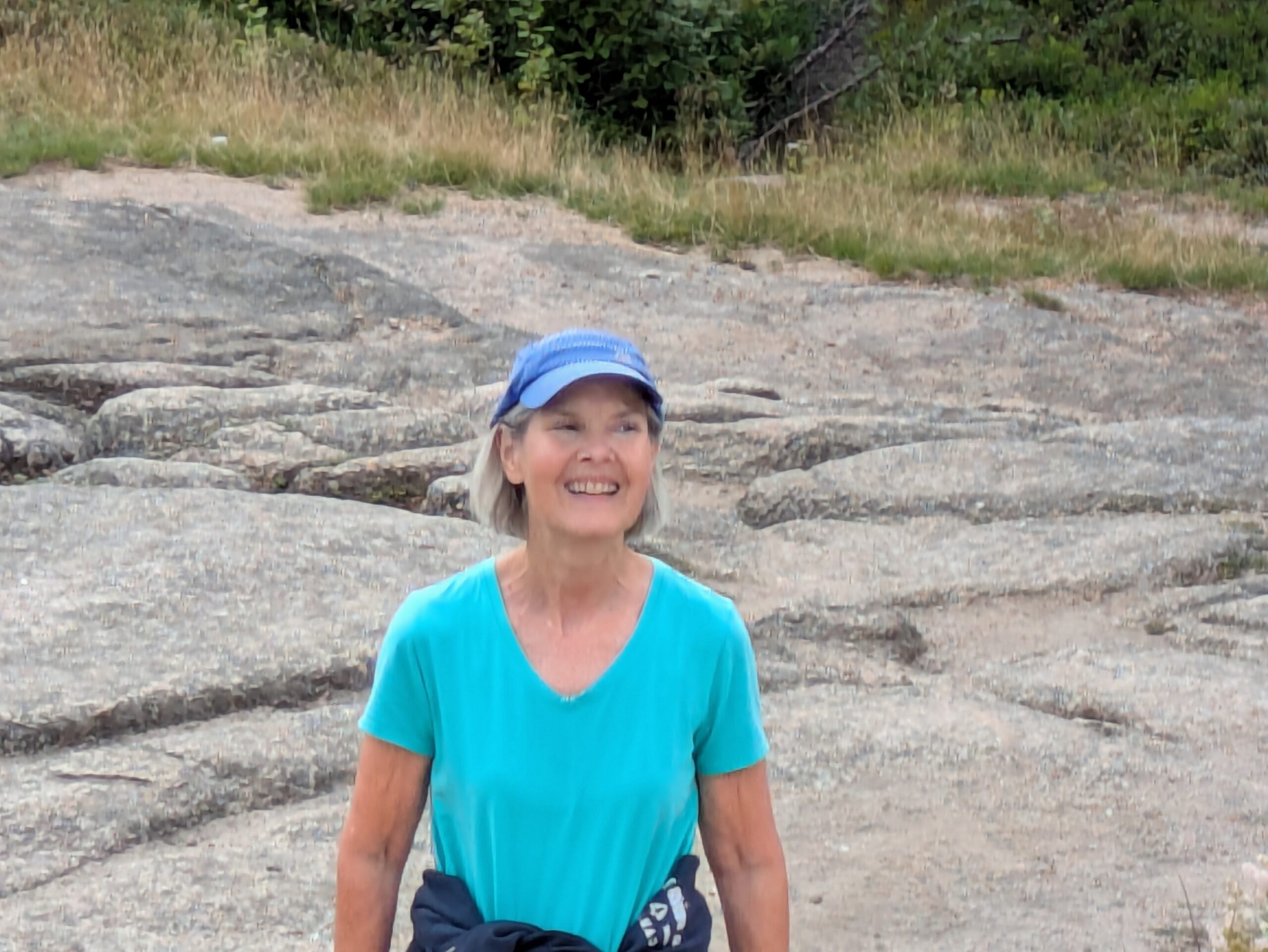 A woman with shoulder length blonde hair standing at the top of a mountain and smiling. She's wearing a blue visor, and a blue t-shirt.