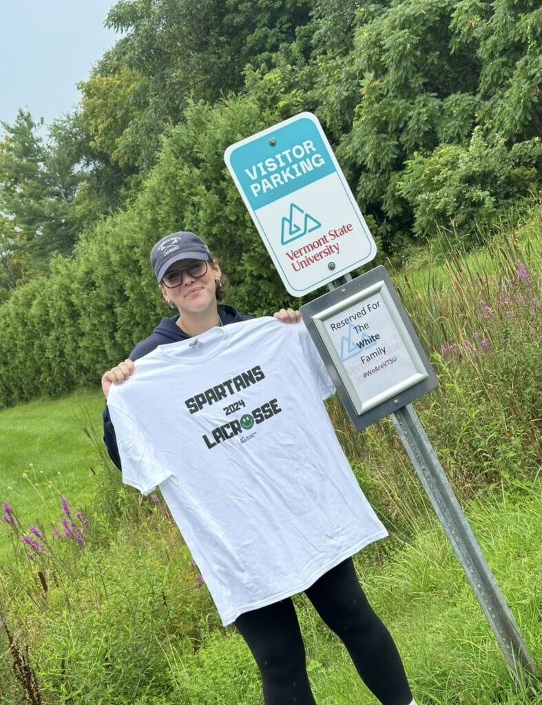 Sam White standing with a white "Spartans Lacrosse" T-shirt held up to her chest. She's standing next to a VTSU Parking sign that reads "Reserved for the White Family"
