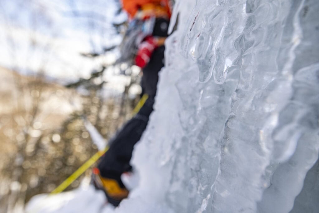 An icy cliff is in focus in the foreground, the legs and torso of an ice climber wearing an orange jacket are blurred in the background