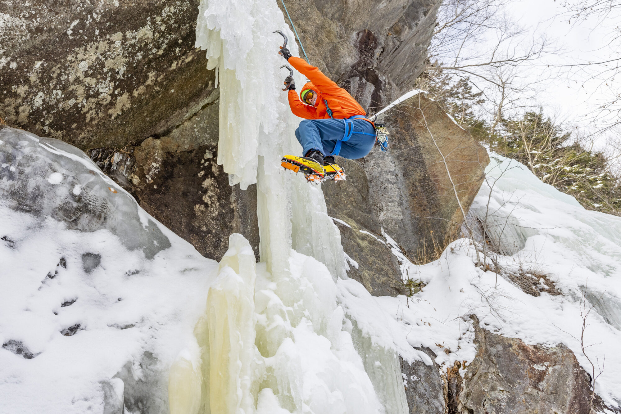 A man wearing an orange jacket and blue pants climbing an icy cliff.