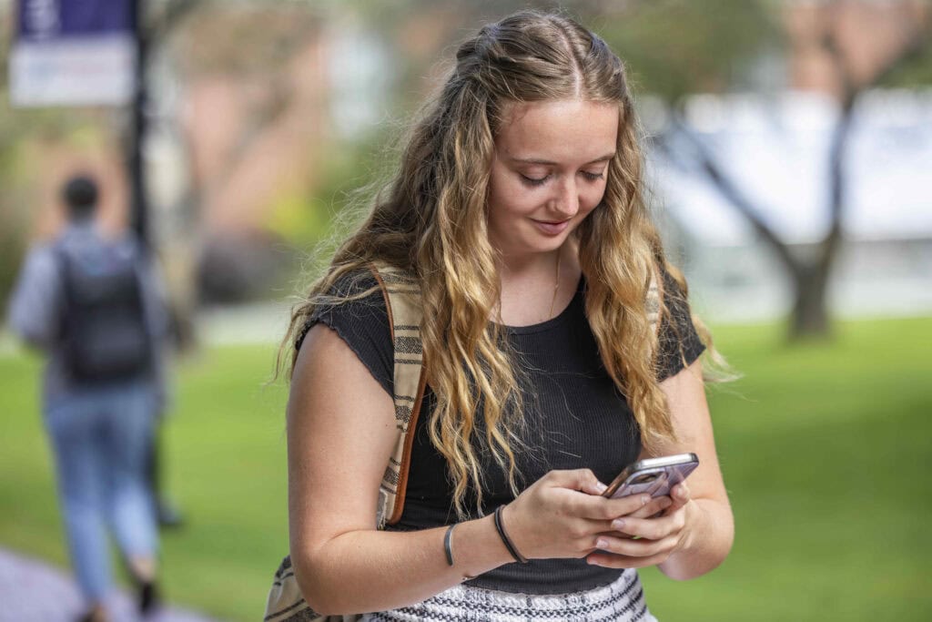 A young woman with long, wavy, blonde hair walking while looking down at her cell phone. She's wearing a black shirt and plaid pants.