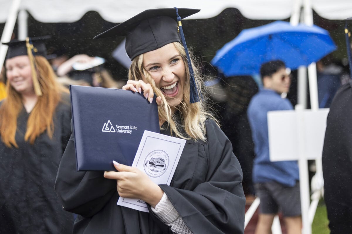 A woman wearing graduation regalia holding up her VTSU Diploma and grinning
