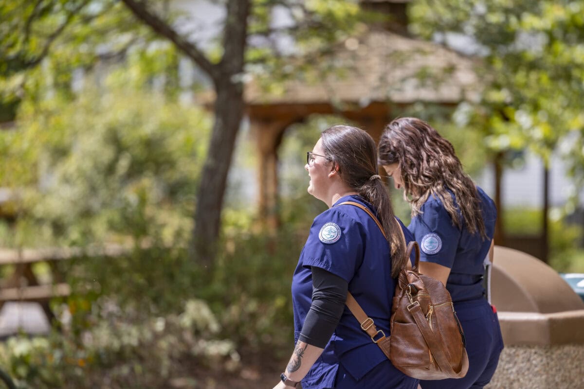 Two female students wearing navy blue scrubs with VTSU Nursing patches on them walk on a path through campus.