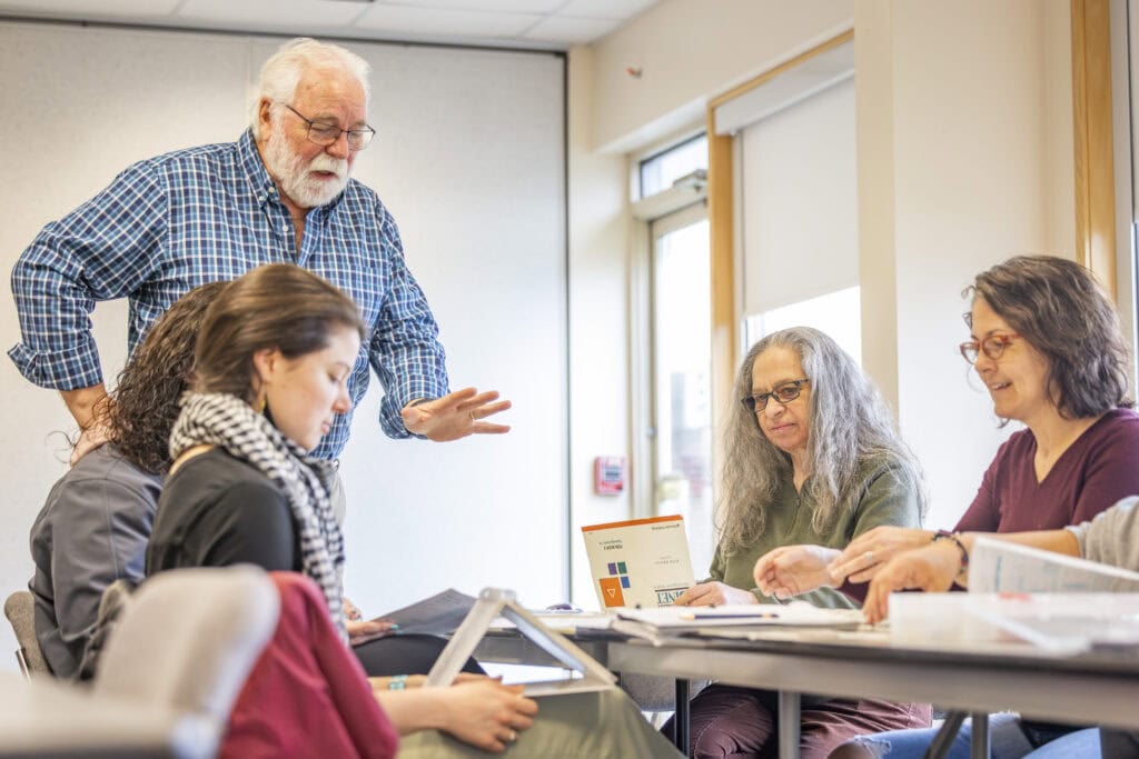 Three students in the Clinical Mental Health program sit at a table during class while their professor is standing and gesturing while he speaks.