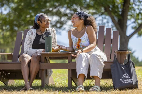 Two students sitting together in wooden chairs chatting in the sunshine.