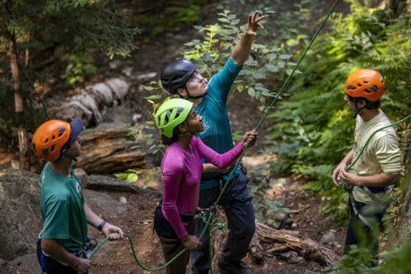 Three students wearing rock climbing gear in the woods being instructed by professor Ben Mirkin, who is pointing upward.
