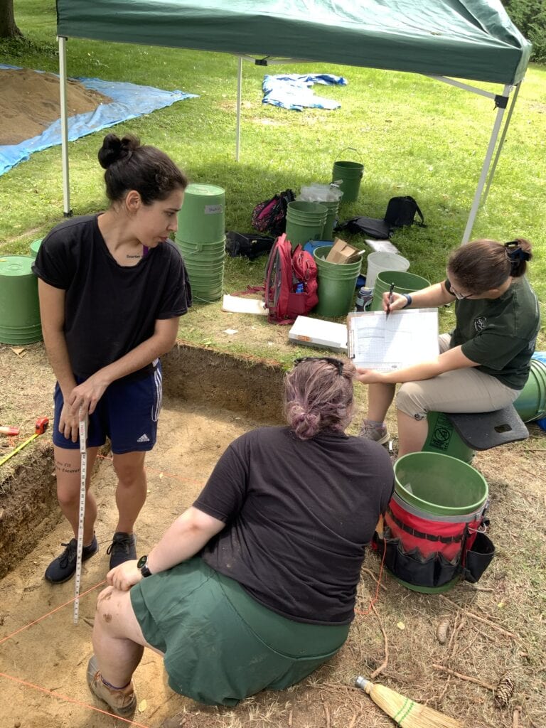 Four people work at an archaeological site beneath a canopy tent. One holds a measuring tape in a soil pit, another writes on a clipboard, while the others assist with the dig. Buckets and tools are scattered around.