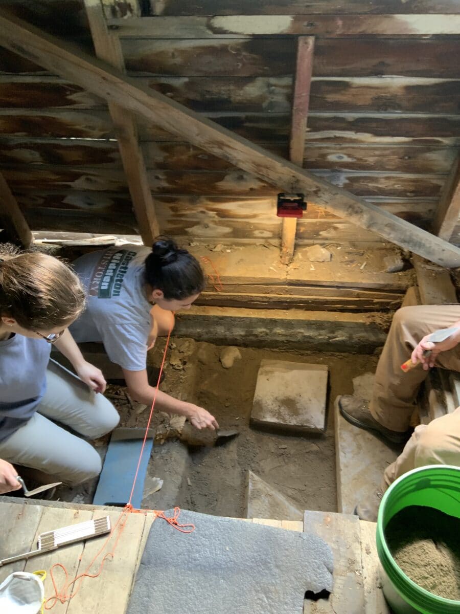 Three individuals kneel inside a wooden structure, carefully excavating soil with small tools. A green bucket filled with dirt sits nearby, and a string grid is set up to guide the dig.