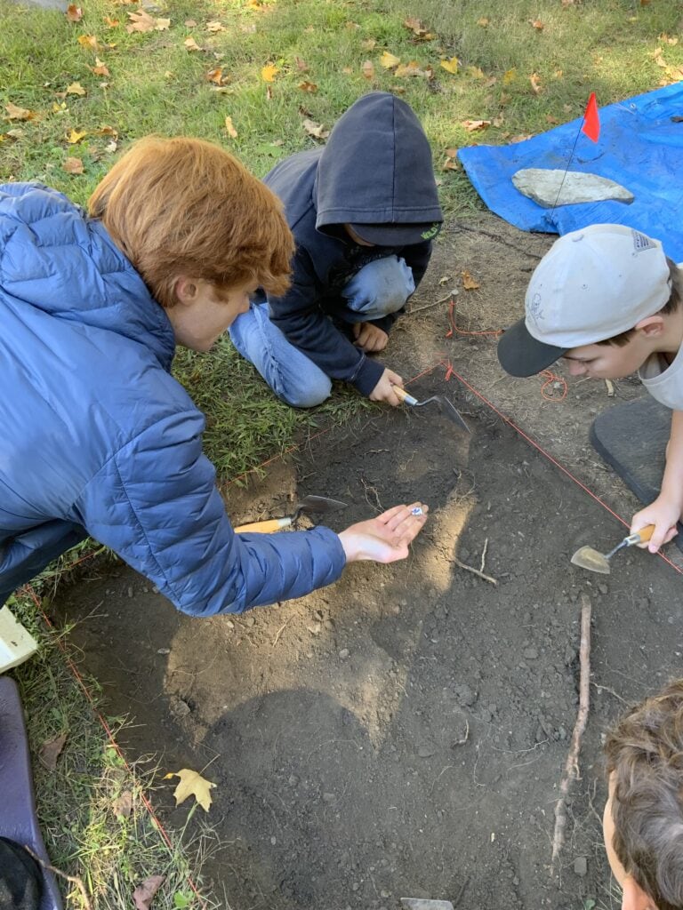 Four individuals conduct a detailed excavation within a square plot marked by red string. They use trowels to carefully dig and examine soil for artifacts.