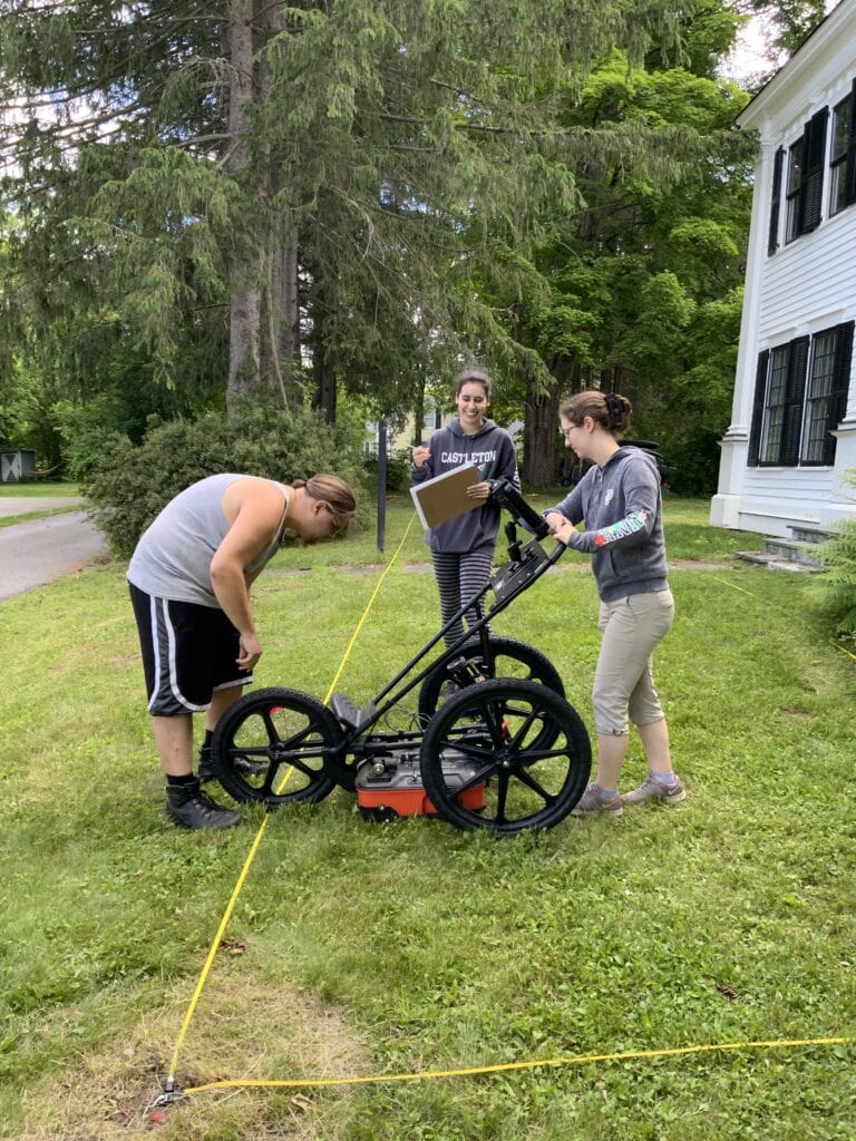 Three individuals operate a wheeled ground-penetrating radar device on a grassy area near trees and houses. One person takes notes on a clipboard as they examine the equipment.