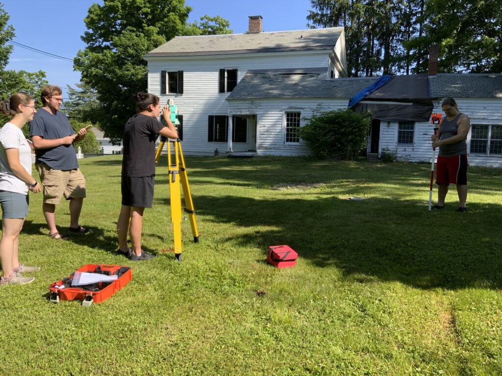 Five people stand or kneel on grass in front of a weathered white house, setting up surveying equipment including tripods and measuring tools.