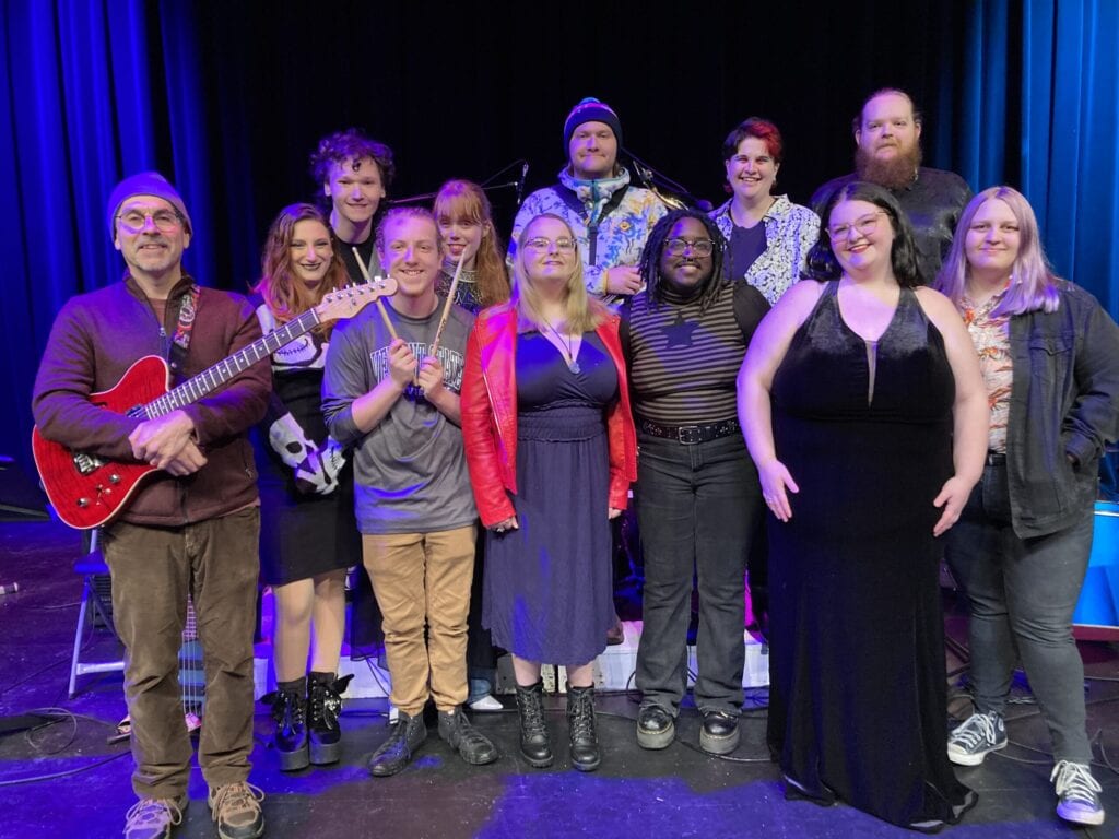 Members of the fall 2024 Funk Fusion ensemble posing for a group photo on stage, with blue light shining on them.