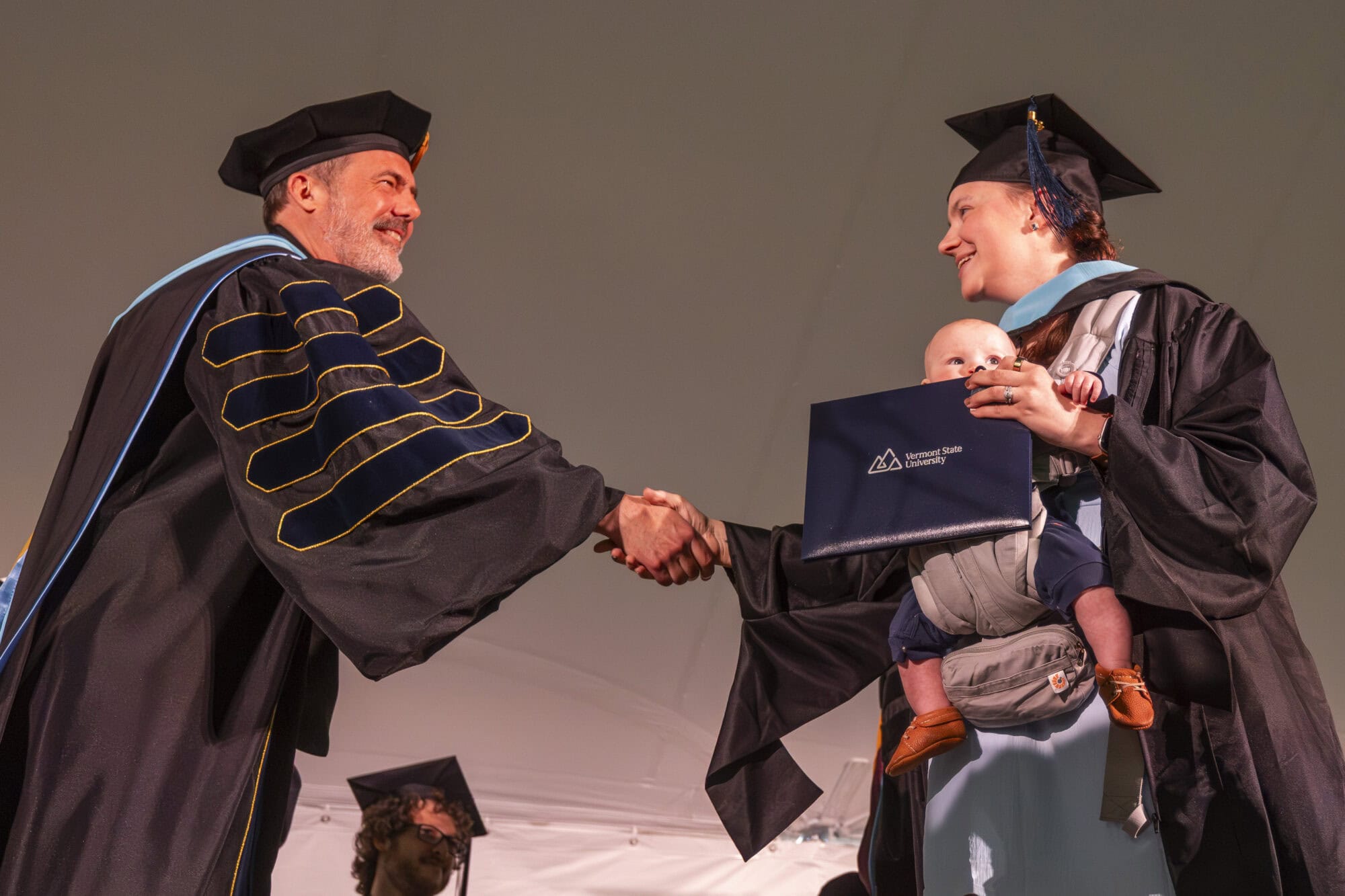 Graduate in academic regalia shakes hands with university president while holding a diploma and carrying a baby in a front carrier during a commencement ceremony.