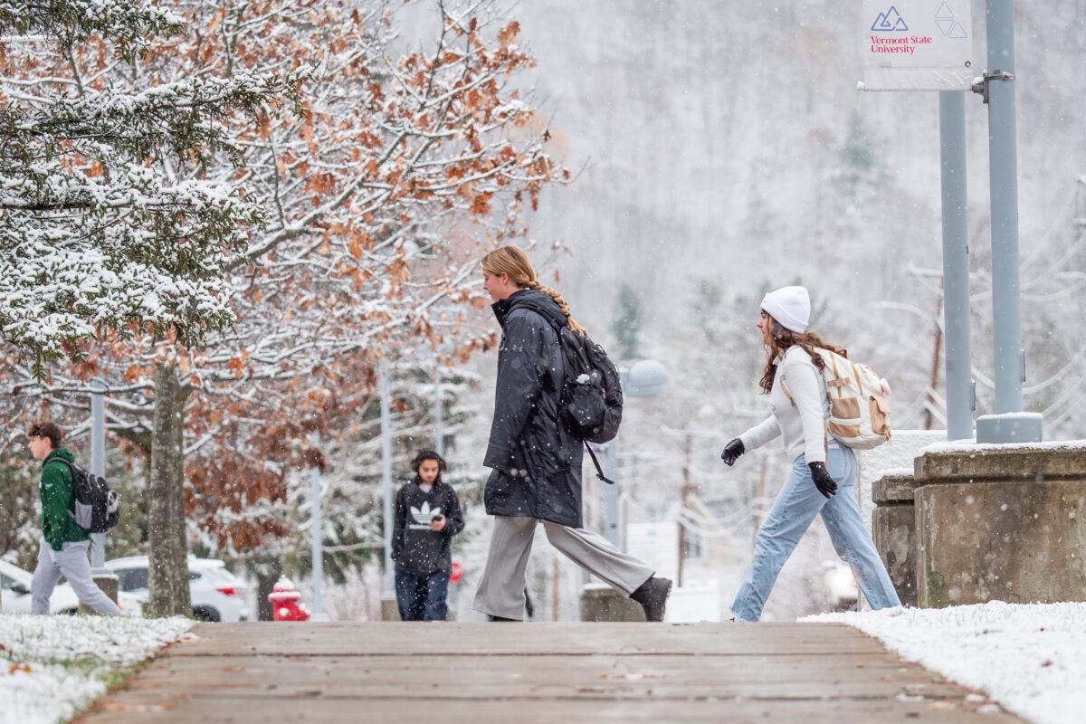 “Students walking across a snow-covered campus sidewalk during the first snowfall of the season. Trees with light snow on branches and a Vermont State University sign are visible in the background.