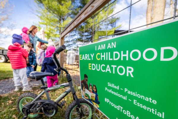 A group of young children dressed in colorful winter clothing stand near a wire fence outdoors, accompanied by an adult. In the foreground, a small bicycle leans against the fence next to a bright green sign that reads: “I AM AN EARLY CHILDHOOD EDUCATOR. Skilled ~ Passionate ~ Professional ~ Essential #ProudToBeAEducator.” Tall trees and a building are visible in the background under a clear sky.
