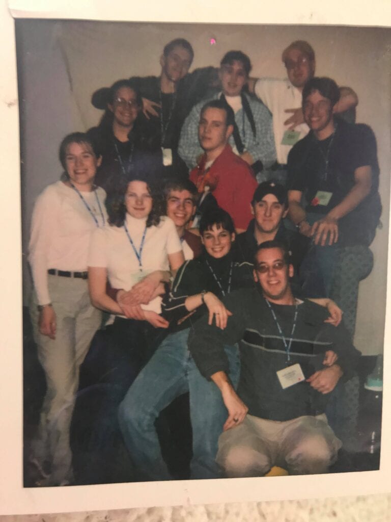 A group of people gathered indoors, sitting and standing closely together in multiple rows. Several individuals are wearing lanyards with name tags. The background is plain, and the group appears to be posing for a casual photo, with some people leaning on others.