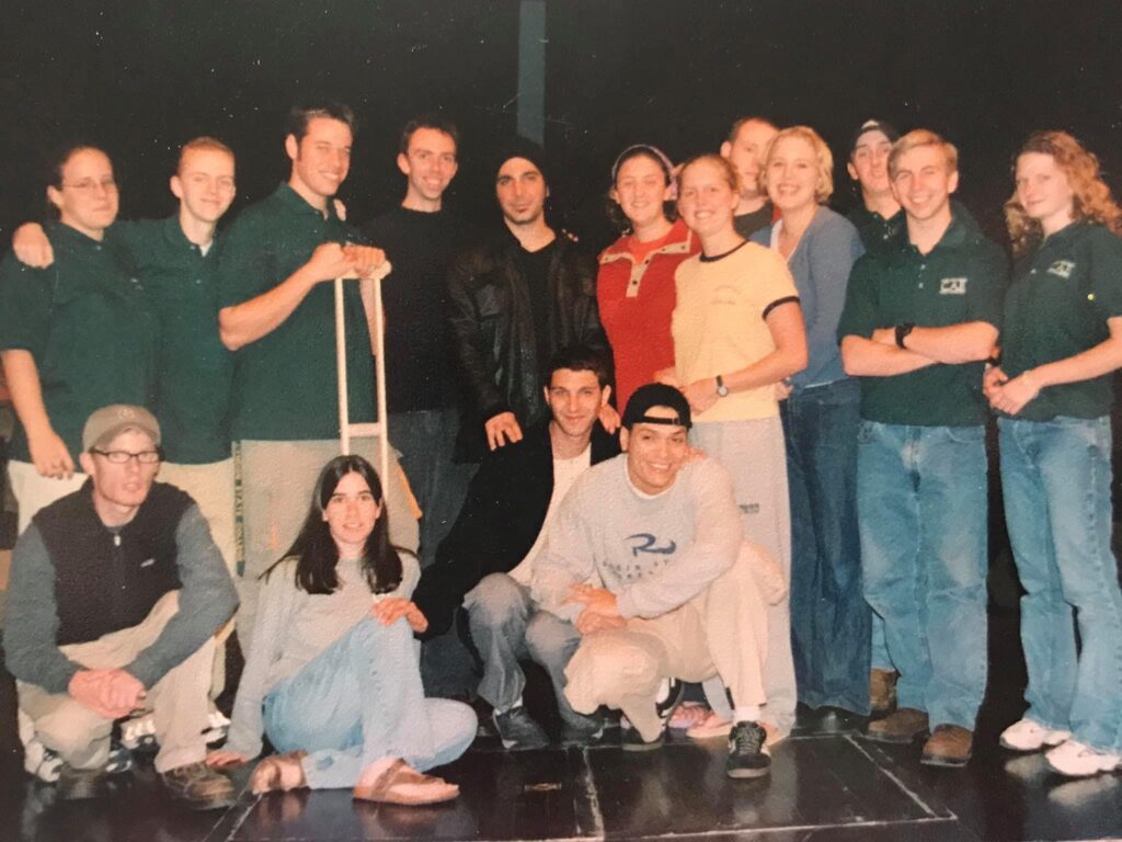 A large group of people posing together indoors on a stage or platform. Most are wearing casual clothing, including jeans and T-shirts, and some are in matching green shirts. Two individuals are seated on the floor in front, and one person is holding crutches. The background is dark, suggesting an auditorium or event space.