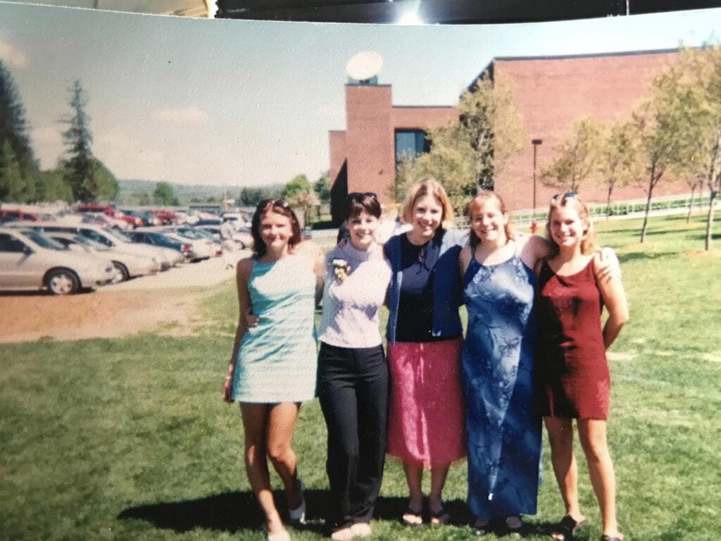 Five people standing outdoors on a grassy area in front of a brick building with parked cars visible in the background. They are dressed in colorful dresses and skirts, suggesting a formal or celebratory occasion. Trees and a clear sky are visible in the background.