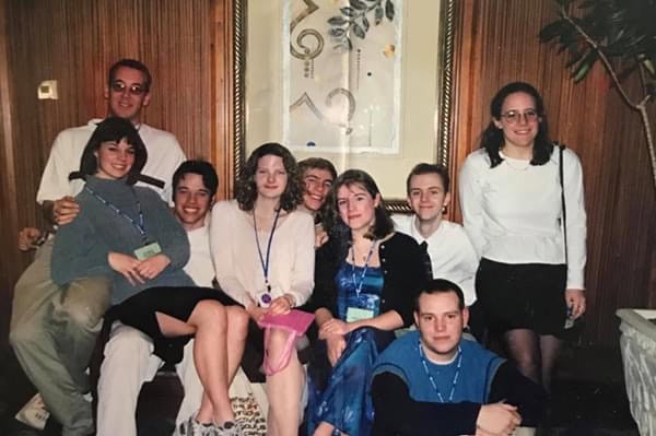 Nine people posing indoors in front of a framed artwork on a wooden-paneled wall. Some are seated on a couch while others stand behind. Several individuals are wearing lanyards with name tags, and the group is dressed in semi-formal attire, including skirts and sweaters.