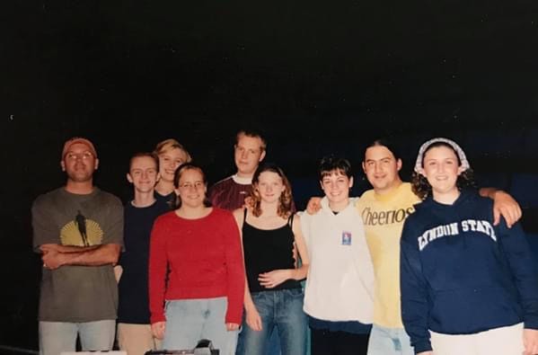 Eight people standing in a row outdoors at night. They are dressed casually in jeans and T-shirts, with one shirt featuring the word “Cheerios” and another reading “Lyndon State.” The group is standing close together, with arms around each other.
