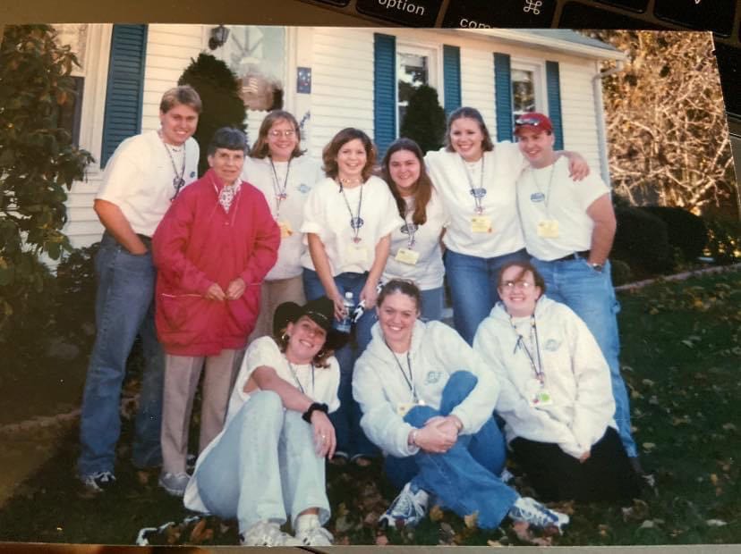 A group of nine people standing and sitting in front of a white house with blue shutters. Most are wearing white shirts with lanyards and name tags, while one person in the center is wearing a bright red jacket. Two individuals are seated on the ground in front, and the rest are standing close together on a lawn covered with fallen leaves.
