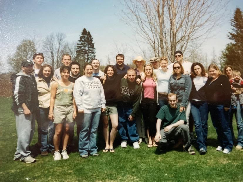 A large group of people standing outdoors on a grassy area with trees in the background. The group is dressed casually in shorts, jeans, and sweatshirts, with one sweatshirt reading “Lyndon State.” The setting appears to be during spring or summer, as the trees have sparse leaves and the grass is green.
