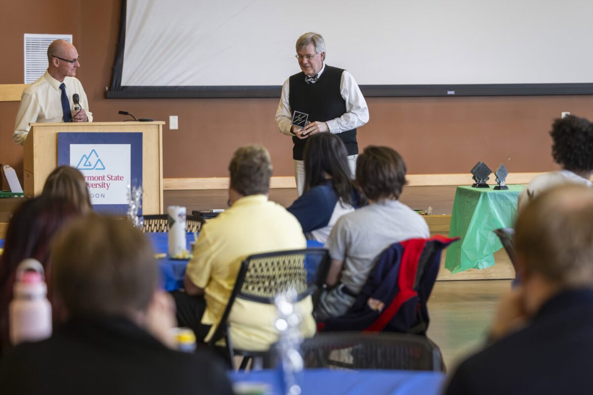 Indoor event at Vermont State University with attendees seated at tables facing a podium and a speaker. A person at the podium holds a microphone, while another person stands near a large screen holding an award. A green table in the background displays additional awards.