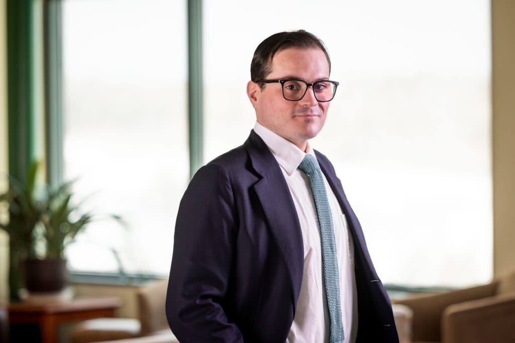 Ernie Fernandez, a man with short brown hair, is wearing rectangular glasses, a black suit, and a grey tie, smiling at the camera
