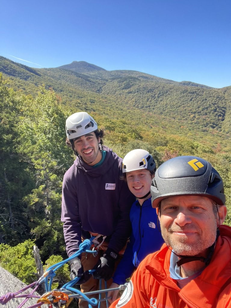Three individuals wearing climbing helmets and harnesses on a rocky ledge with ropes secured. Behind them is a scenic view of forested mountains under a clear blue sky.