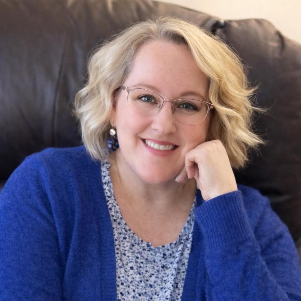 Airen Wallen, a woman with short, wavy, blonde hair is wearing a blue and white shirt with a blue sweater over top, with round glasses. She is smiling widely at the camera.
