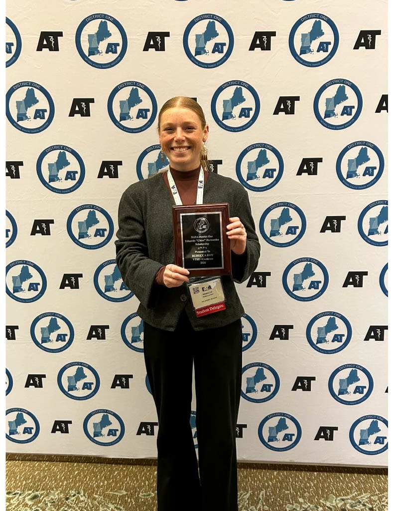 A student stands in front of an Eastern Athletic Trainers’ Association step‑and‑repeat banner holding a plaque award and wearing conference credentials.
