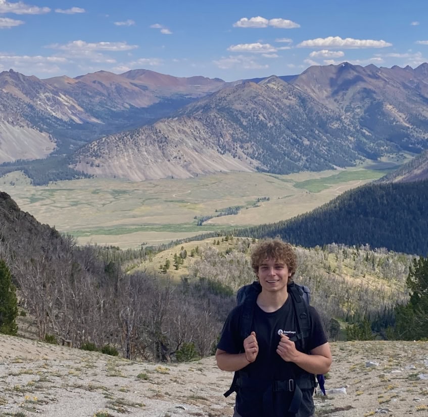 Ben Boggio, a young man with curly blonde hair is standing atop a mountain with a sweeping mountain range in the background