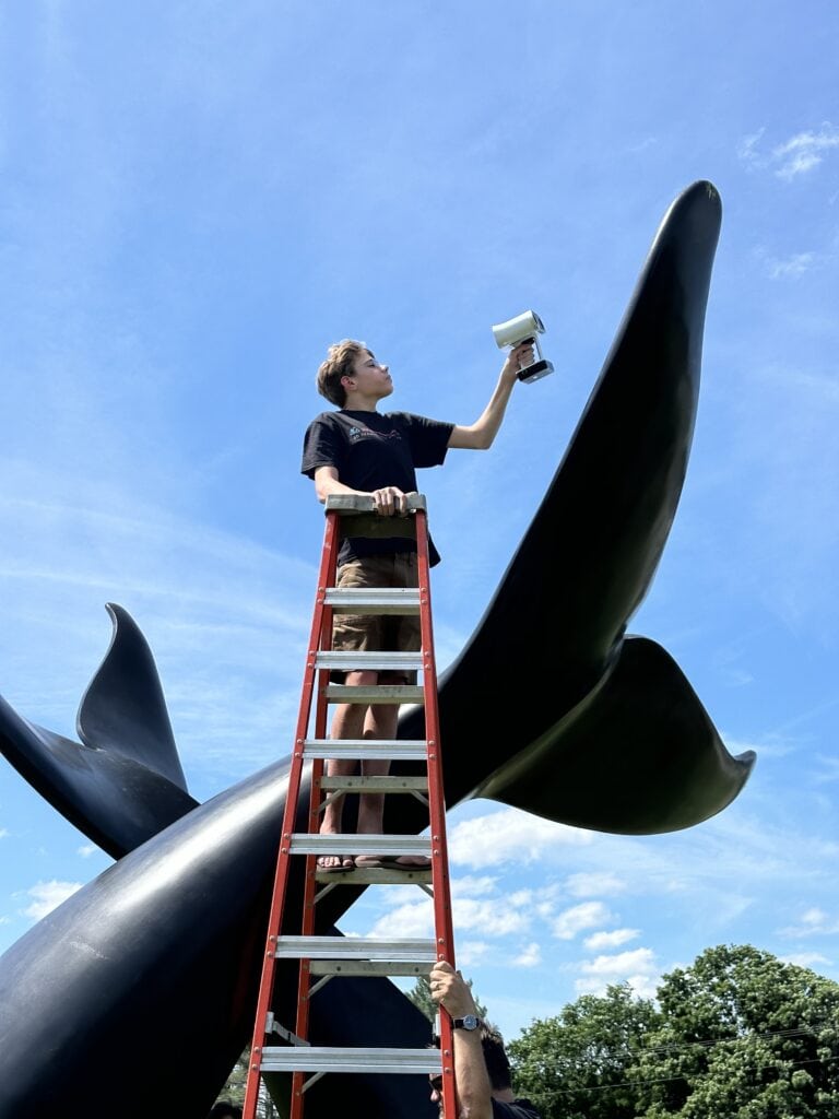 A student at the top of a ladder using 3D imaging tools to scan the Whale Tails sculpture in Randolph vt