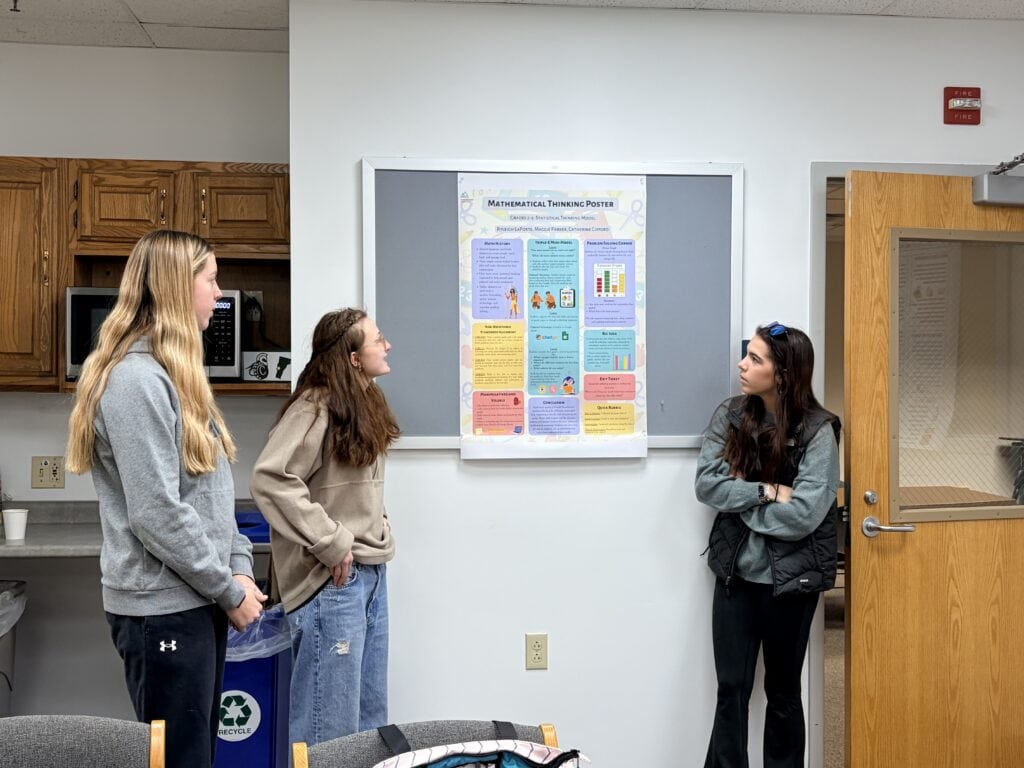 Three students standing in front of a poster titled “Measurement Thinking Poster.” The poster includes sections on measurement concepts and strategies, with colorful graphics. The setting includes a classroom with a recycling bin and a door.
