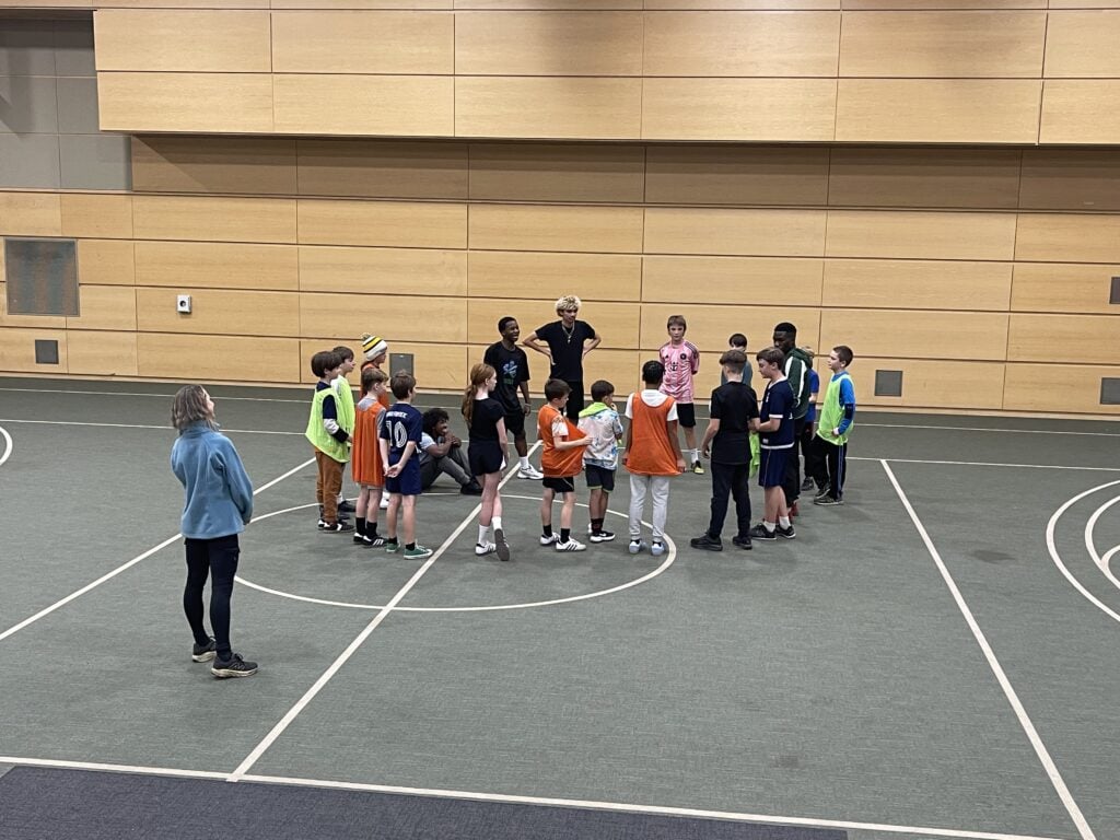 A large circle of children gathers at mid‑court in the gym as student‑athlete coaches stand in the center giving instructions.
