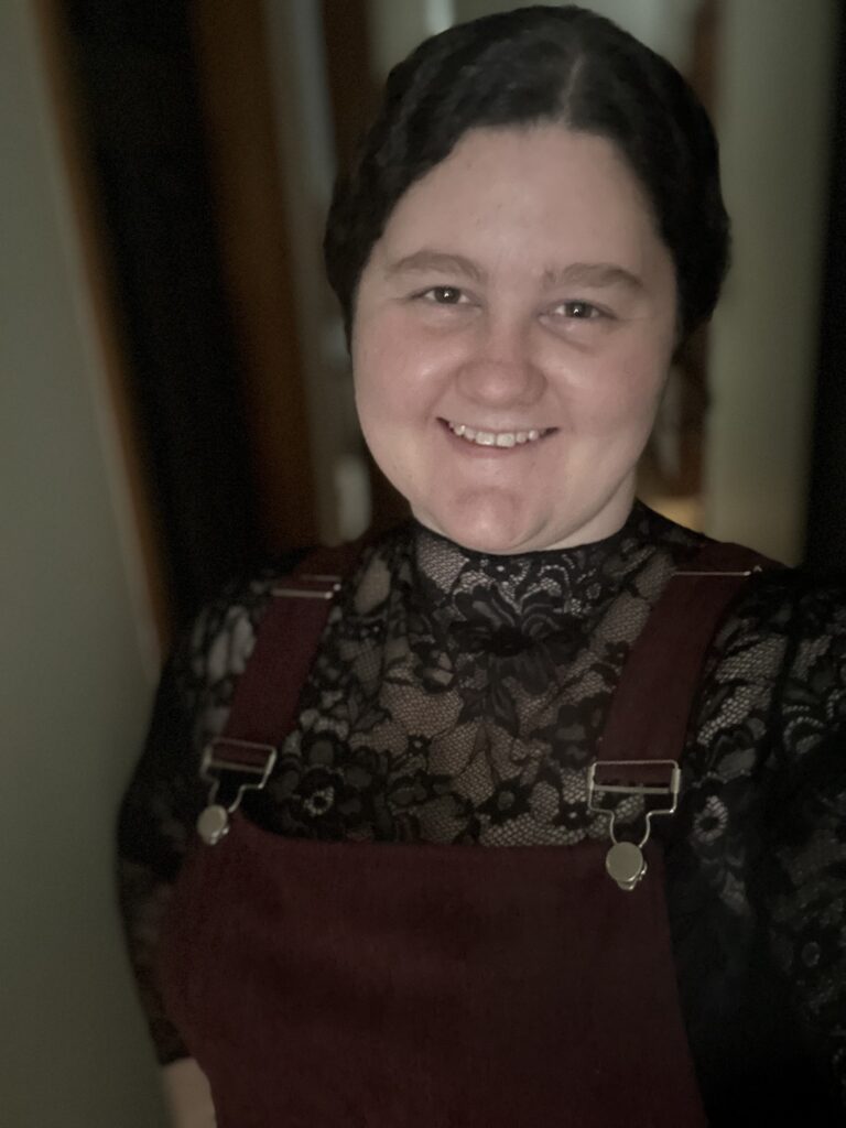 Jerika LaValley, a woman with brown hair tied in a ponytail and wearing black overalls over a black patterned shirt, smiling at the camera.