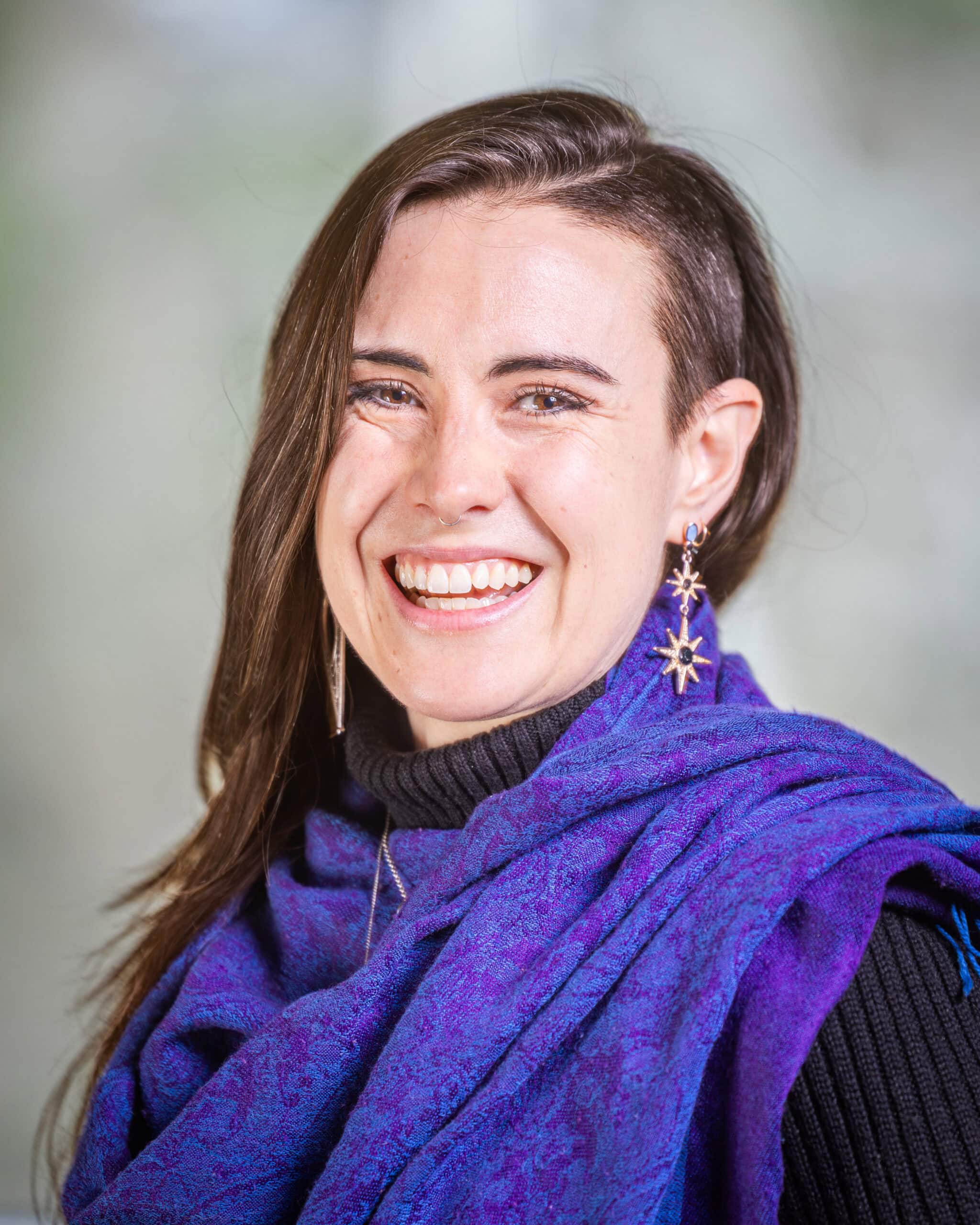 Joy Haqq, a person with long brown hair is wearing a purple scarf over a black turtleneck and smiling at the camera.