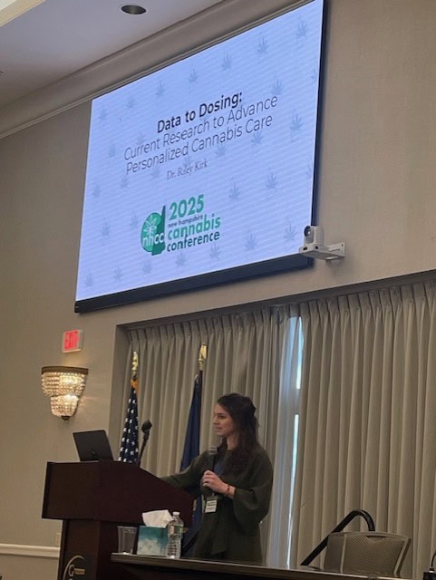 A speaker stands at a podium in a conference room presenting under a large screen displaying the title “Data to Dosing: Current Research to Advance Personalized Cannabis Care” by Dr. Riley Kirk. The screen also shows the 2025 NH Cannabis Conference logo. The podium has a microphone and a laptop, and the background includes curtains and U.S. flags.