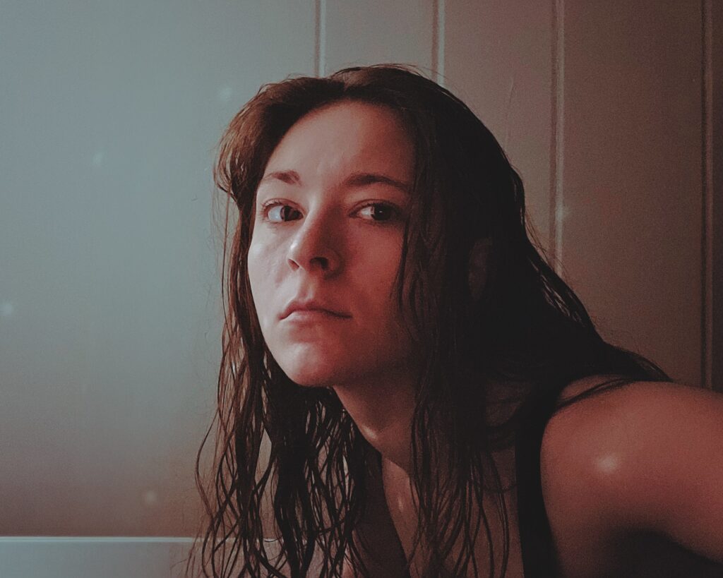 Rebecca Kutcher, a woman with long brown hair is leaning forward on her elbows with white tiles in the background.