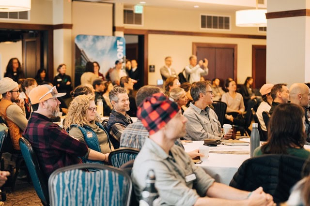 A large group of attendees seated at round tables in a conference room, listening to a presentation. The room has warm lighting, wood-paneled doors, and a banner in the background with the word “Vermont.”