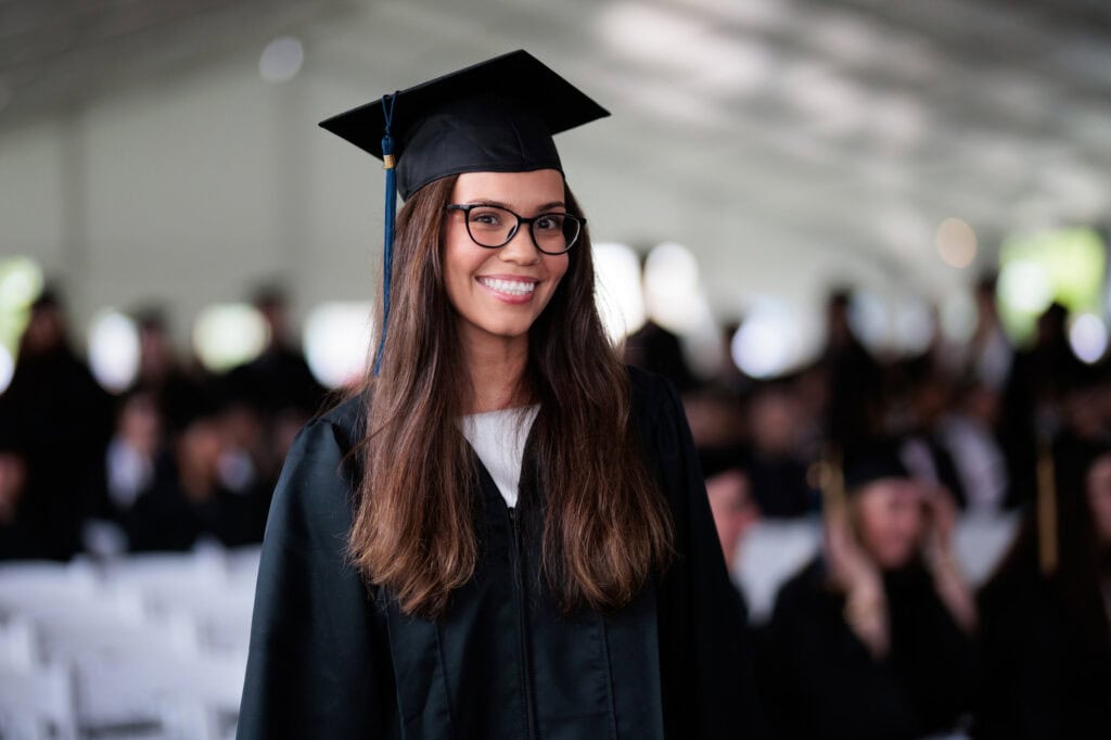 A young woman with long brown hair and round glasses smiling at the camera while wearing graduation regalia
