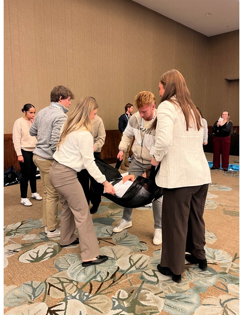 Students participate in a hands‑on workshop, practicing a scenario by lifting a peer in a soft stretcher during a training exercise.
