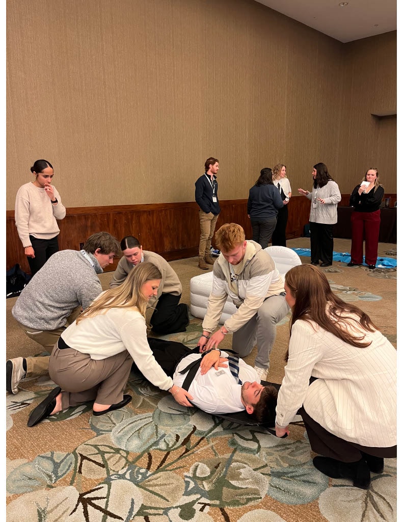 A group of students kneel on the floor practicing an emergency care scenario, working together to stabilize a peer lying on the ground during a workshop.
