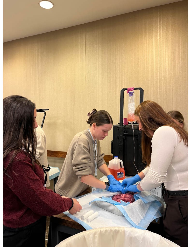 Students gather around a table during a medical workshop, wearing gloves and handling training materials on a protective pad.
