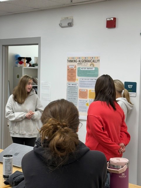 Three students standing near a wall-mounted poster titled “Thinking Algebraically!”. The poster contains sections on standards, history, and problem-solving strategies. The students appear to be engaged in discussion about the content.