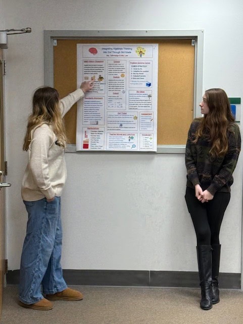 Two students standing in front of a bulletin board displaying a poster titled “Integrating Algebraic Thinking Into 2nd Through 3rd Grade.” The poster includes math history, problem-solving strategies, and standards alignment.
