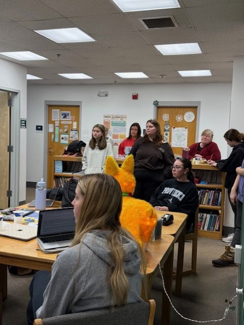A classroom setting with multiple students gathered around tables and standing near walls. The room has bookshelves, posters, and a door with educational materials displayed. A bright orange plush animal is on one of the tables next to laptops and water bottles. The environment appears collaborative and informal.
